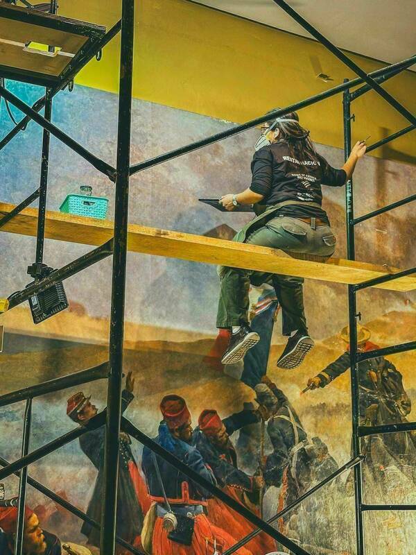 A person wearing a black t-shirt, khaki pants, and a face mask sits on scaffolding while restoring a large mural. They hold a palette and brush, carefully working on the upper section of the painting. Below, the mural depicts a vivid battle scene with soldiers on horseback, engaging in combat.