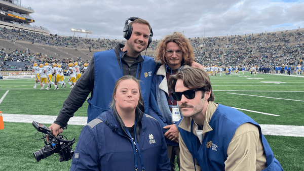 Four Notre Dame media personnel pose for a photo on the sidelines of Notre Dame Stadium during a football game.  They are wearing navy blue vests with the Fighting Irish Media logo.  In the background, the football teams are on the field.
