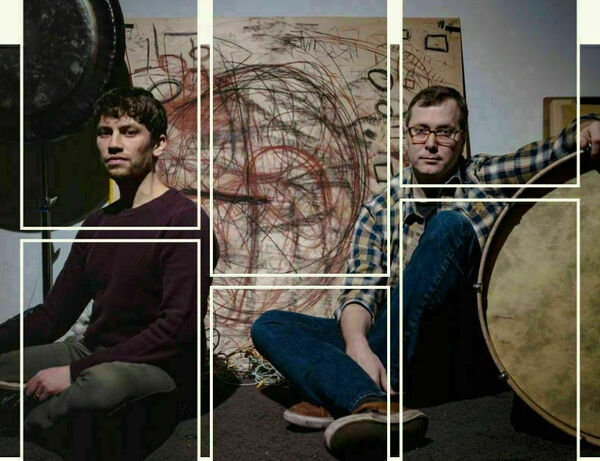 Two men sit on a dark floor near a large artwork of red and black scribbles and a large circular drum. One man wears a dark purple sweater and olive green pants; the other, glasses, a plaid shirt, and jeans.