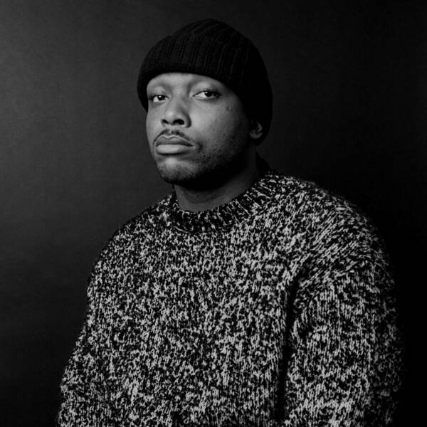 Black and white headshot of a person wearing a dark knit beanie and a speckled black and white sweater, looking directly at the camera with a serious expression against a dark background.