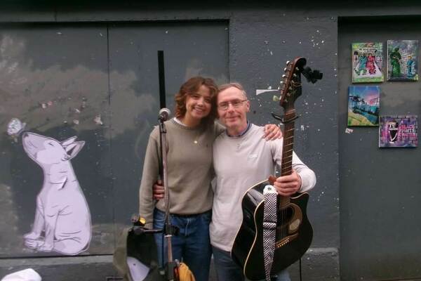 Two people stand against a gray wall with art and posters. A young woman with shoulder-length brown hair, wearing an olive sweater and jeans, smiles at the camera with her arm around a man.  He holds an acoustic guitar and wears glasses and a gray long-sleeved shirt.