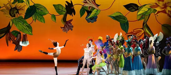 A dancer in a light brown deer costume with antlers and dark eye makeup performs an arabesque. Behind them, a diverse group of dancers in colorful, elaborate costumes with feather headpieces, skeleton masks, and flowing skirts stand against a vibrant orange backdrop with large painted flowers.