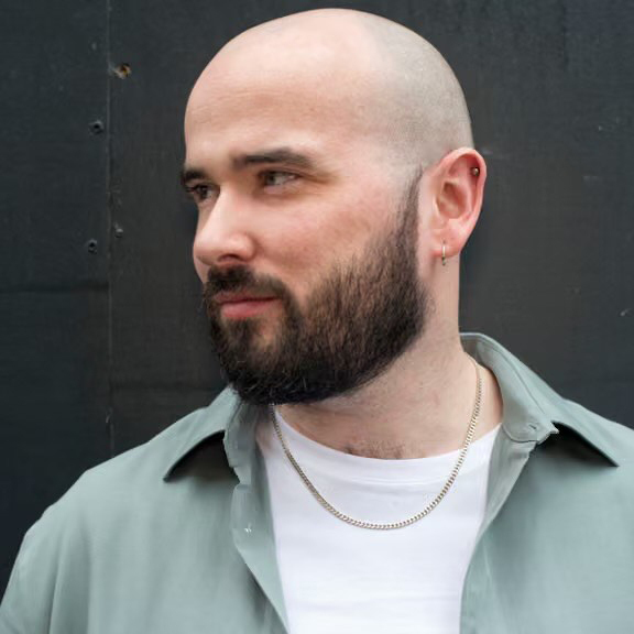 A bald man with a full dark beard and light complexion looks to his left. He wears a light green collared shirt over a white t-shirt, a silver chain, and ear piercings.