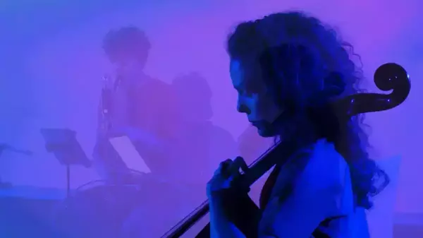 A person with dark, curly hair and a headset mic plays a cello, focused under vibrant blue and purple lighting. Another musician, indistinctly visible in the blurry background, plays a wind instrument.