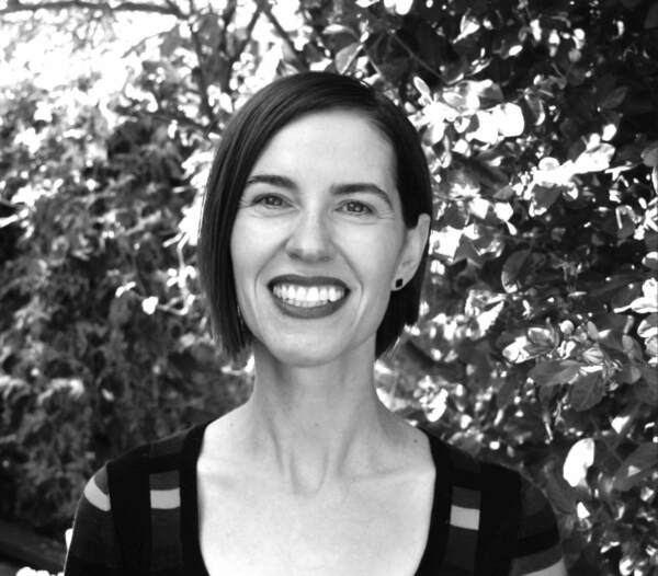 Smiling woman with short dark hair, dark lipstick, and a striped top, against blurred foliage.