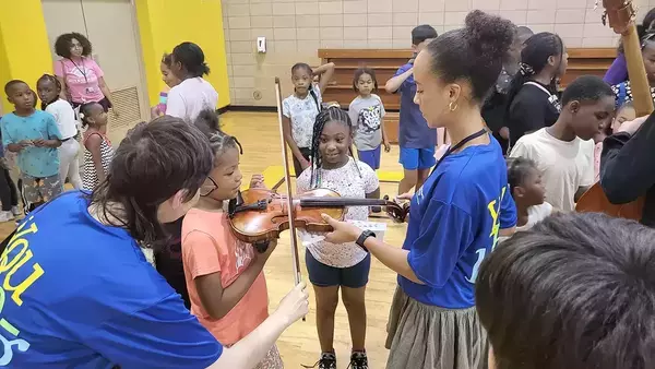 Two adults help young girls in a bright gymnasium learn to play violins, smiling as they hold the instruments. Other children observe.