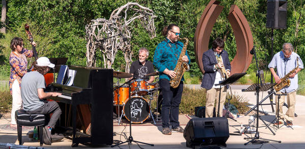 Six musicians play outdoors: piano, upright bass, drums, two saxophones, and trumpet. Green trees and sculptures in background.