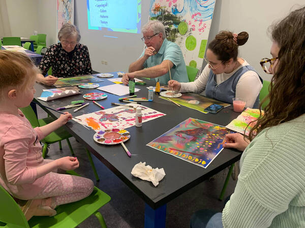 Diverse group of people, from child to senior, painting vibrant artwork at a black table in a bright art workshop.