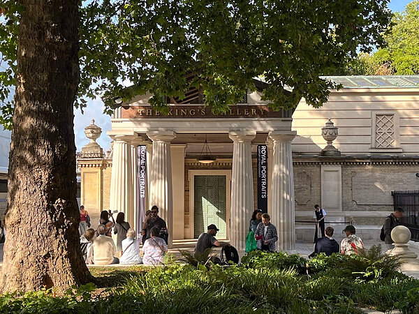 The King's Gallery building with white columns and "ROYAL PORTRAITS" banners. Visitors sit under a large tree.