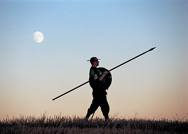 Warrior in helmet and armor holds a round shield and spear, silhouetted on a grassy ridge against a twilight sky with a moon.